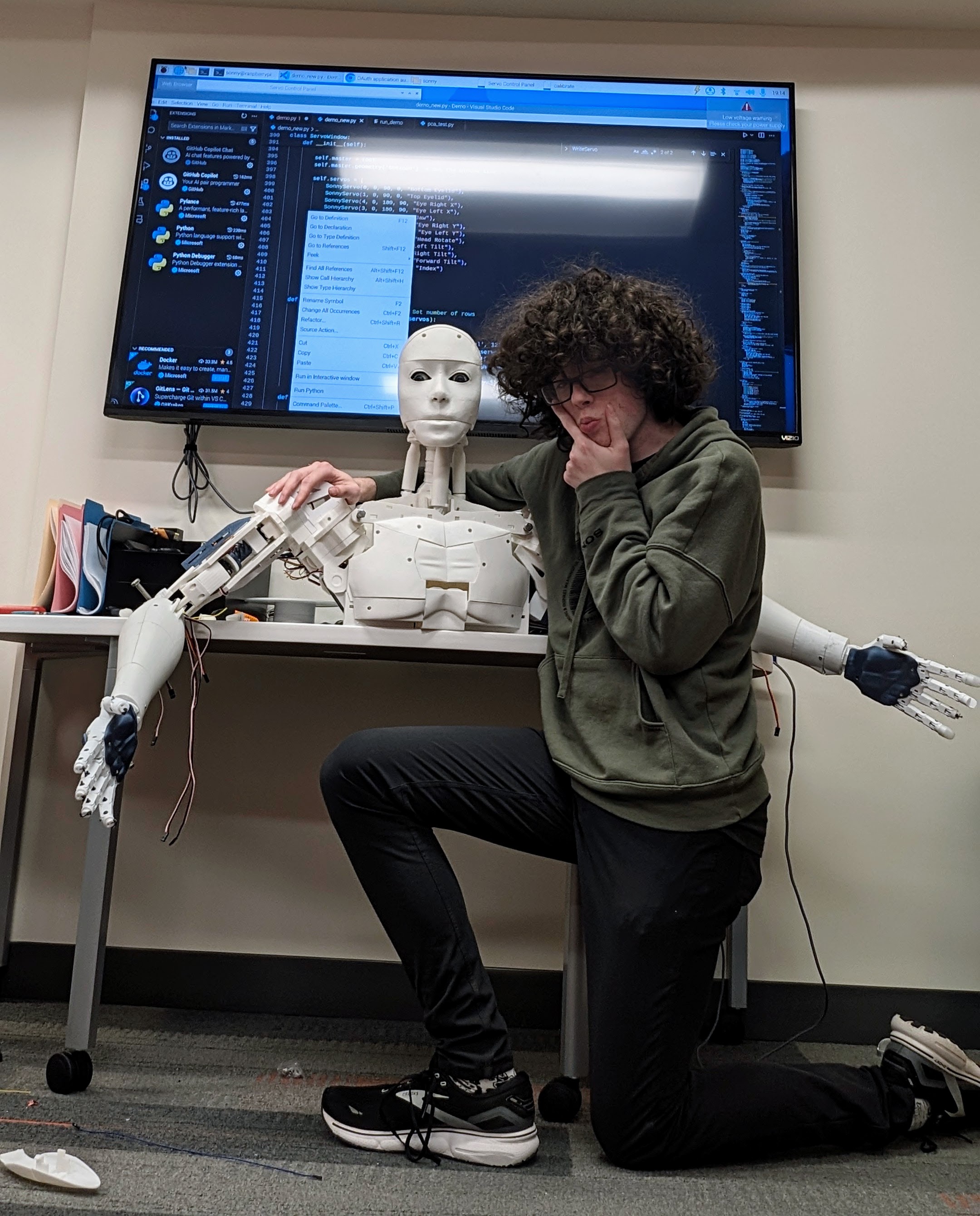 Rio beside the 3D-printed humanoid in the lab, its servo control code on the screen behind.